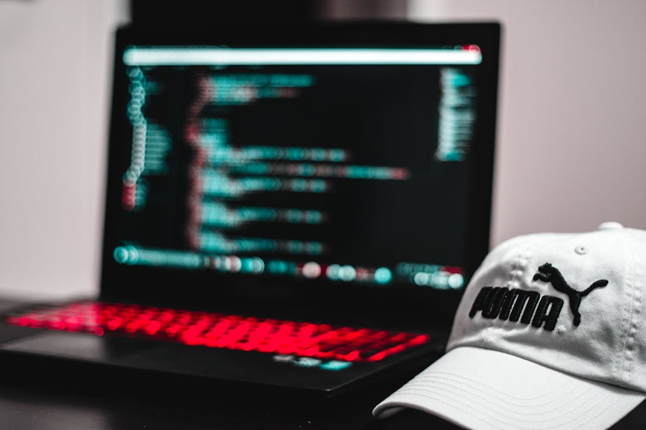 A black laptop with a red-backlit keyboard displaying lines of computer code on a dark screen, positioned on a plain, smooth surface. Next to the laptop, in the foreground, is a white Puma baseball ca